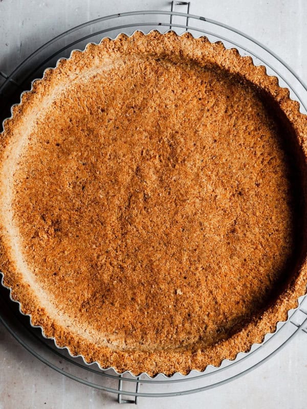 Golden-brown, baked almond flour tart crust in a fluted tart pan placed on a metal cooling rack, set against a light-colored, textured background. The crust has an even, crumbly texture and precise edges, suggesting it is freshly made and ready for filling.