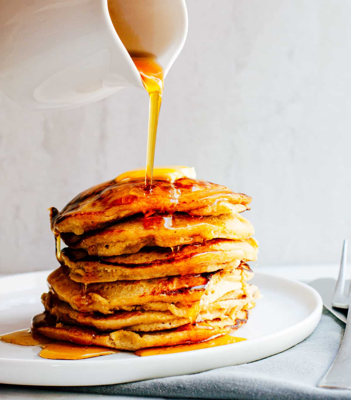 Pouring maple syrup over a stack of Banana Oatmeal Pancakes on a white plate.