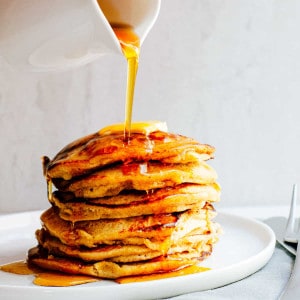 Pouring maple syrup over a stack of Banana Oatmeal Pancakes on a white plate.