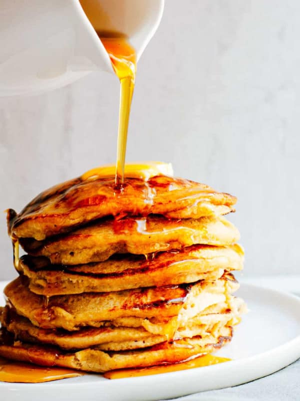 Pouring maple syrup over a stack of Banana Oatmeal Pancakes on a white plate.