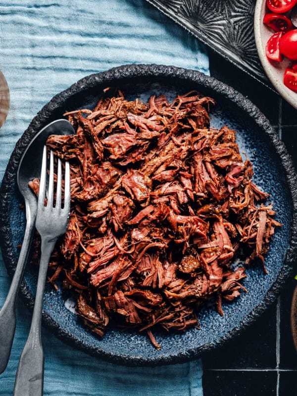 A dark blue plate filled with shredded beef, flanked by a fork and a knife on the left side. Beside the plate are a glass of beer, a bowl of cherry tomatoes, a small dish of lime wedges, and a garnish of fresh herbs on a blue textured surface.