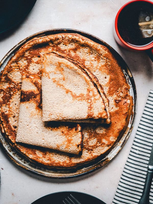 A plate of buckwheat crepes folded in quarters, accompanied by a red mug of tea with a tea bag string hanging out, a knife, and a striped napkin on a light textured table surface.