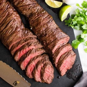 sliced carne asada made in the oven on a black cutting board with knife and citrus fruits photographed from above.