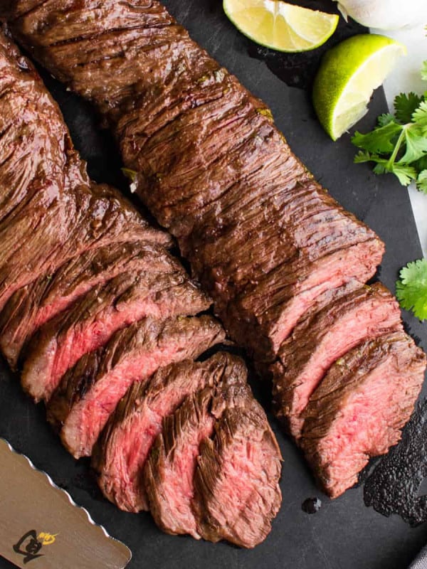 sliced carne asada made in the oven on a black cutting board with knife and citrus fruits photographed from above.