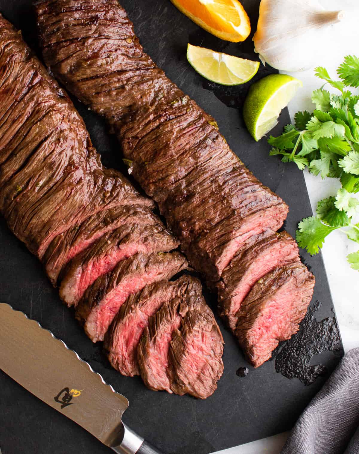 sliced carne asada made in the oven on a black cutting board with knife and citrus fruits photographed from above.