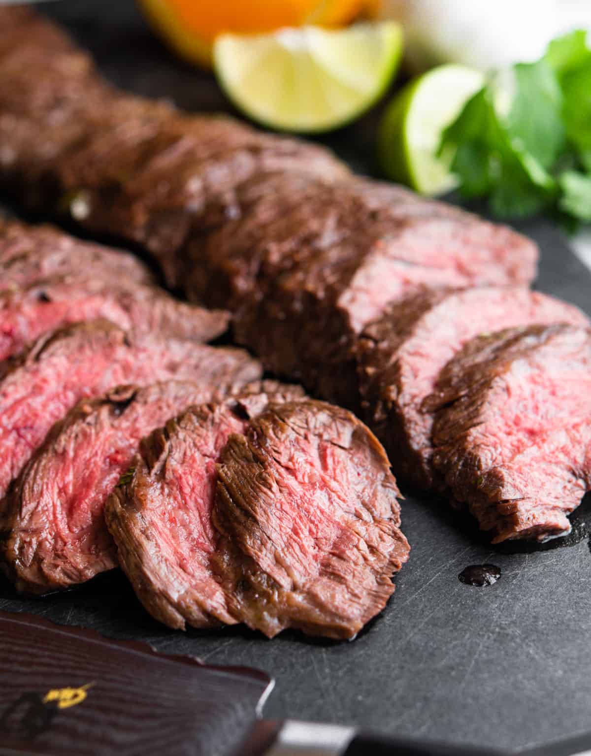 sliced carne asada made in the oven on a black cutting board with knife and citrus fruits.