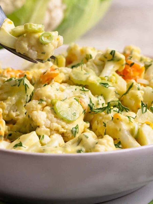 One bite of Cauliflower Potato Salad on a fork from a white serving bowl photographed at an angle.