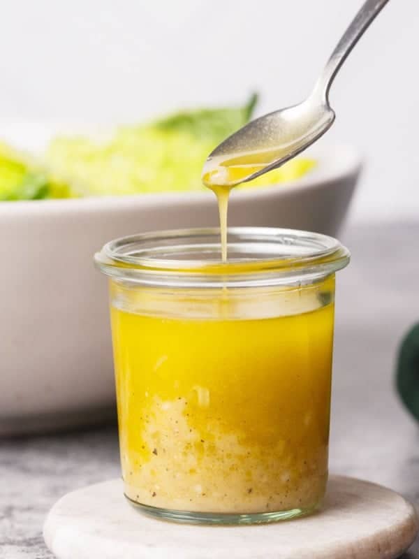 A spoon drizzles an olive oil and vinegar salad dressing into a small glass jar filled with the dressing over a light-colored saucer. A bowl of leafy green salad is blurred in the background, and a dark green cloth is partially visible to the side.