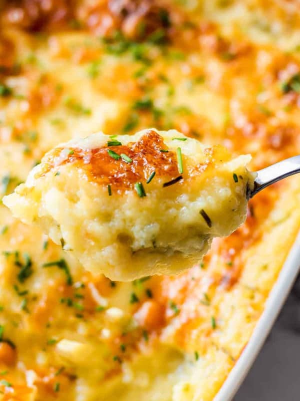 A scoop of Cheesy Mashed Potatoes with a white baking dish full of them below.