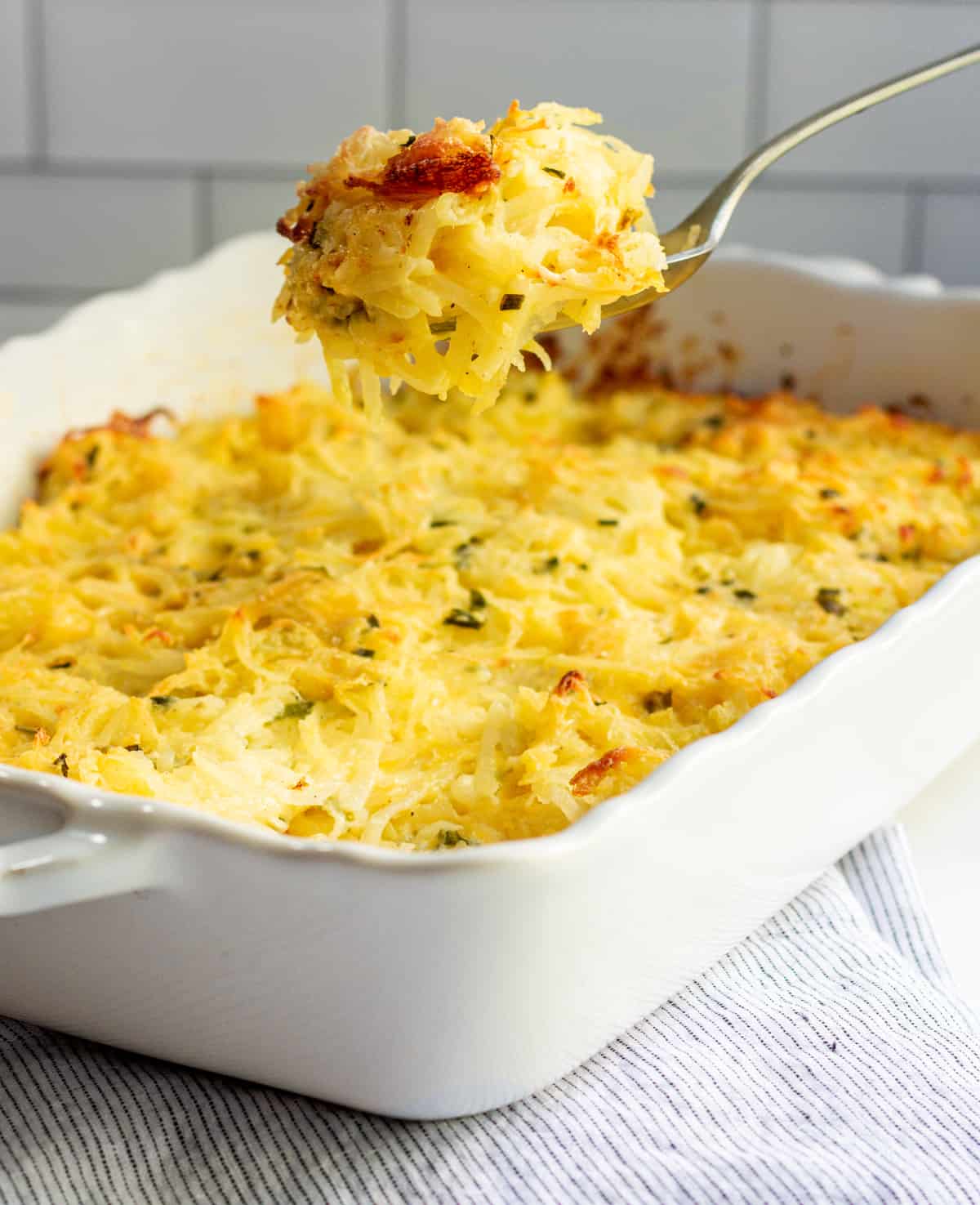 Serving Cheesy Potatoes from a white ceramic baking dish with a silver spoon.