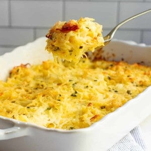 Serving Cheesy Potatoes from a white ceramic baking dish with a silver spoon.