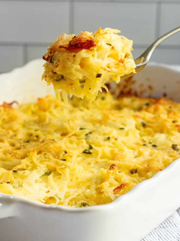 Serving Cheesy Potatoes from a white ceramic baking dish with a silver spoon.