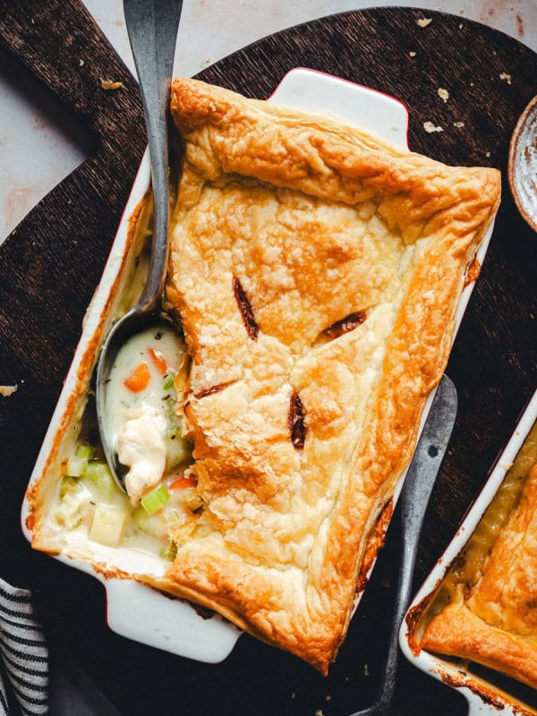 A rectangular baking dish with a golden-brown puff pastry crust, partially removed to reveal a creamy vegetable and chicken filling inside. A spoon rests inside the dish. Surrounding items include a bowl of coarse salt, a small dish with herbs, and a striped cloth napkin.