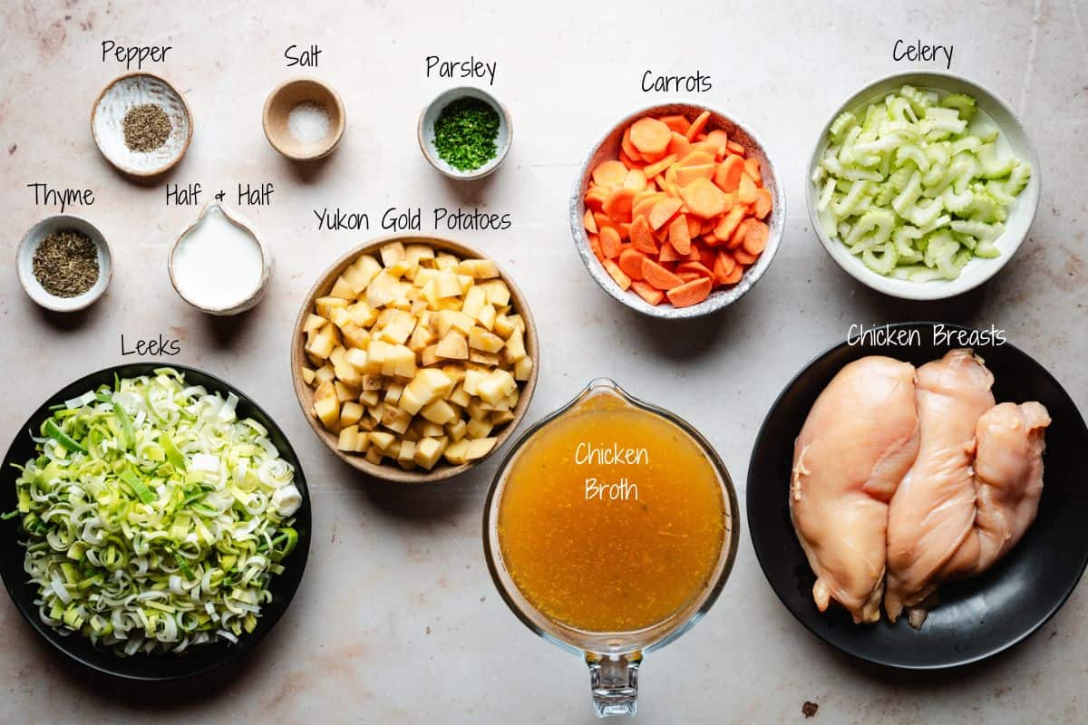 Ingredients for a chicken potato soup arranged on a table: pepper, salt, parsley, thyme, half & half, diced Yukon Gold potatoes, chopped leeks, carrots, celery, chicken breasts, and a jug of chicken broth.