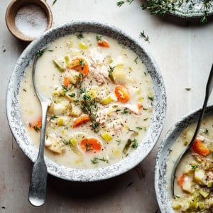 A bowl of creamy chicken soup with vegetables such as carrots and potatoes, garnished with fresh thyme. A spoon rests in the bowl. Another partial bowl, herbs, and a small dish of salt are visible on a light textured surface.