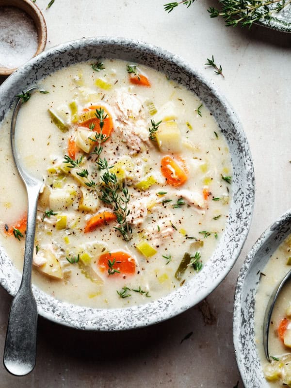 A bowl of creamy chicken soup with vegetables such as carrots and potatoes, garnished with fresh thyme. A spoon rests in the bowl. Another partial bowl, herbs, and a small dish of salt are visible on a light textured surface.