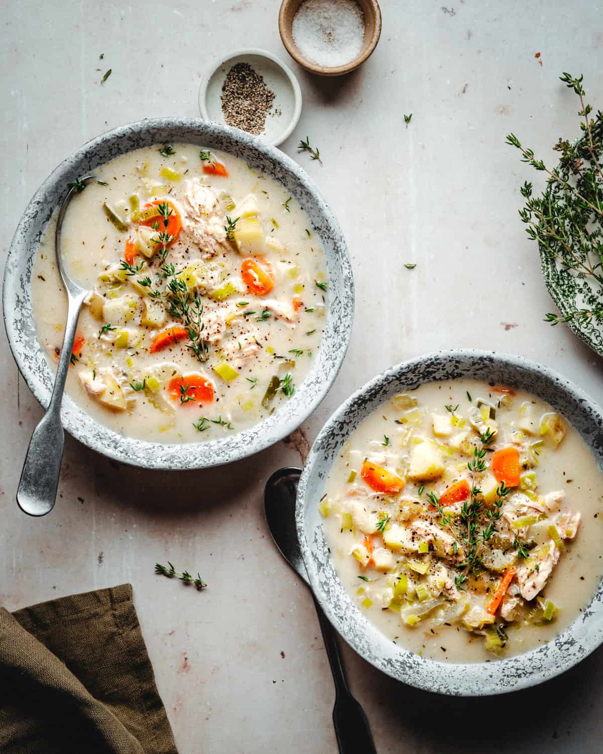 Two bowls of creamy chicken potato soup with carrots, celery, and herbs, garnished with fresh thyme. A spoon rests in one bowl with additional thyme sprigs, salt, and pepper nearby on a light countertop. A green cloth is partially visible.