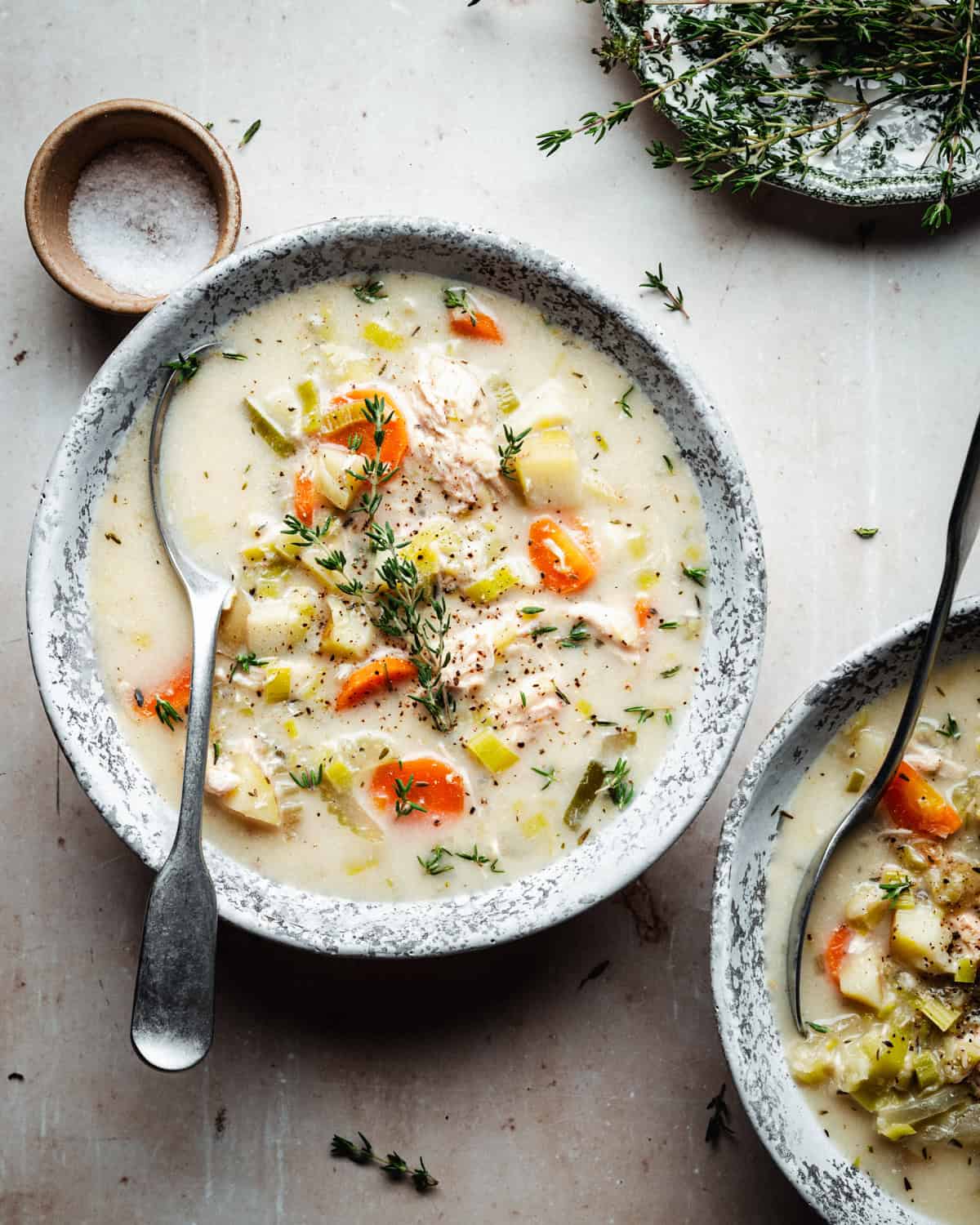 A bowl of creamy chicken and potato soup garnished with fresh thyme. The soup includes carrots, potatoes, and green beans. A spoon rests in the bowl, with a small dish of salt and a sprig of thyme nearby on a light-colored surface.