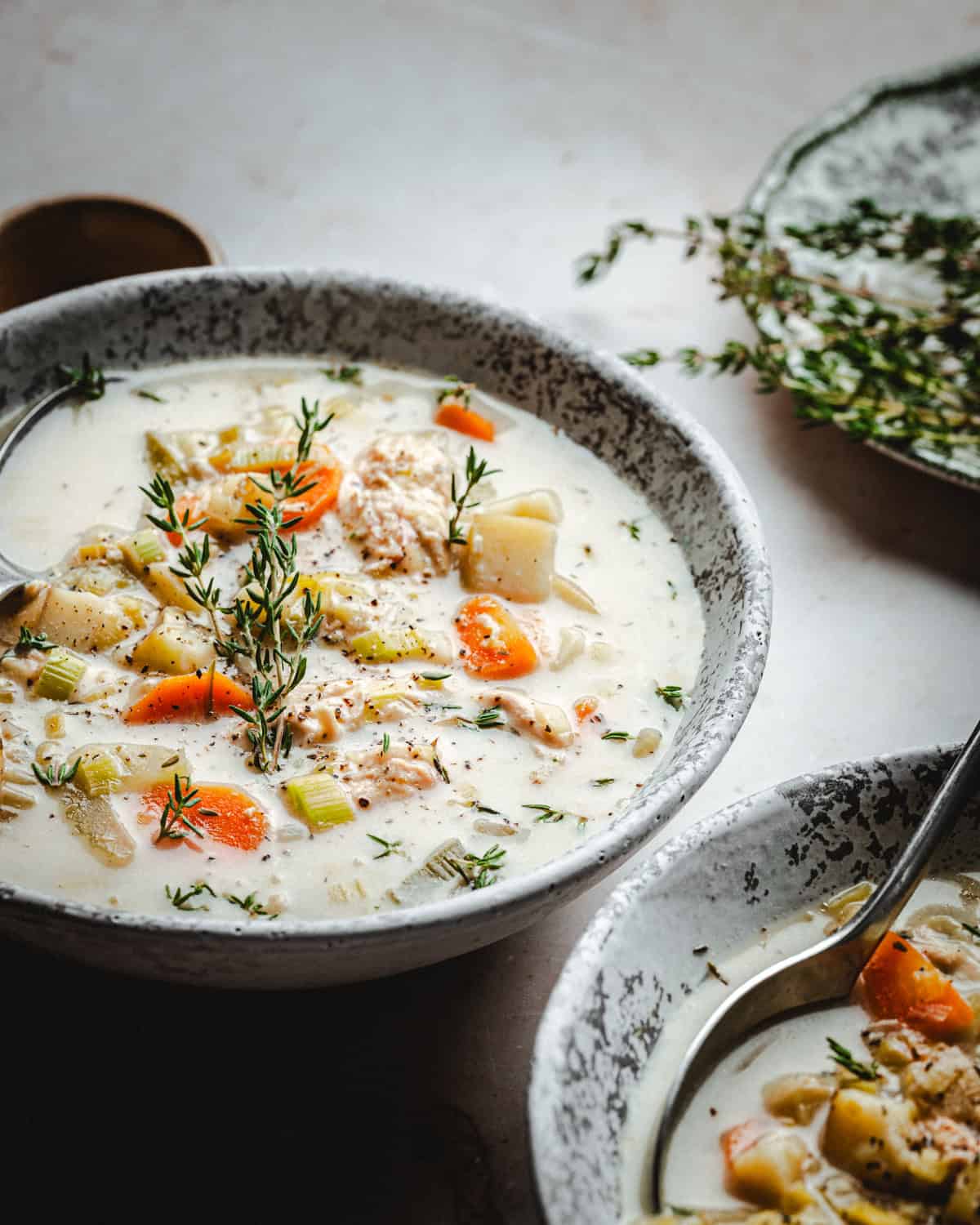 A bowl of creamy chicken potato soup filled with chunks of chicken, carrots, potatoes, and celery, garnished with fresh thyme. A spoon rests in the bowl, and a sprig of thyme lies on the table beside it.