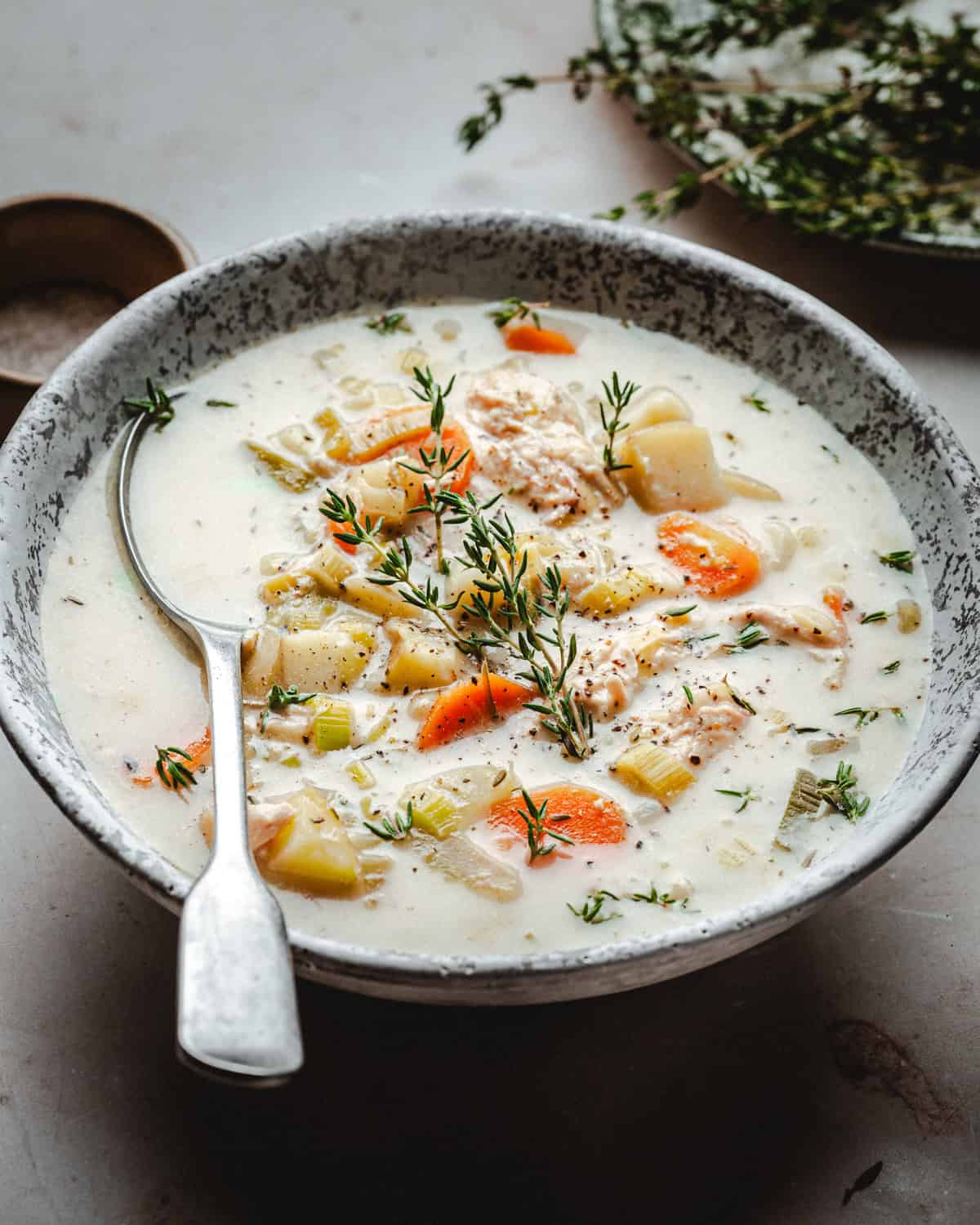 A bowl of creamy chicken and potato soup filled with chunks of vegetables and shredded chicken, garnished with fresh thyme sprigs. A spoon rests in the bowl, and a small dish of herbs is visible in the background.