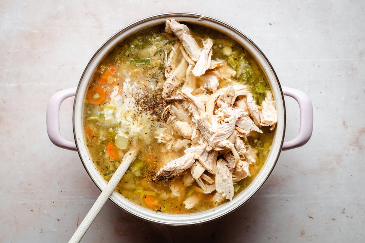 A pot of chicken soup with shredded chicken, sliced carrots, celery, and onions in broth. A wooden spoon rests in the soup, which is seasoned with herbs and spices, visible from a top-down view on a light countertop.