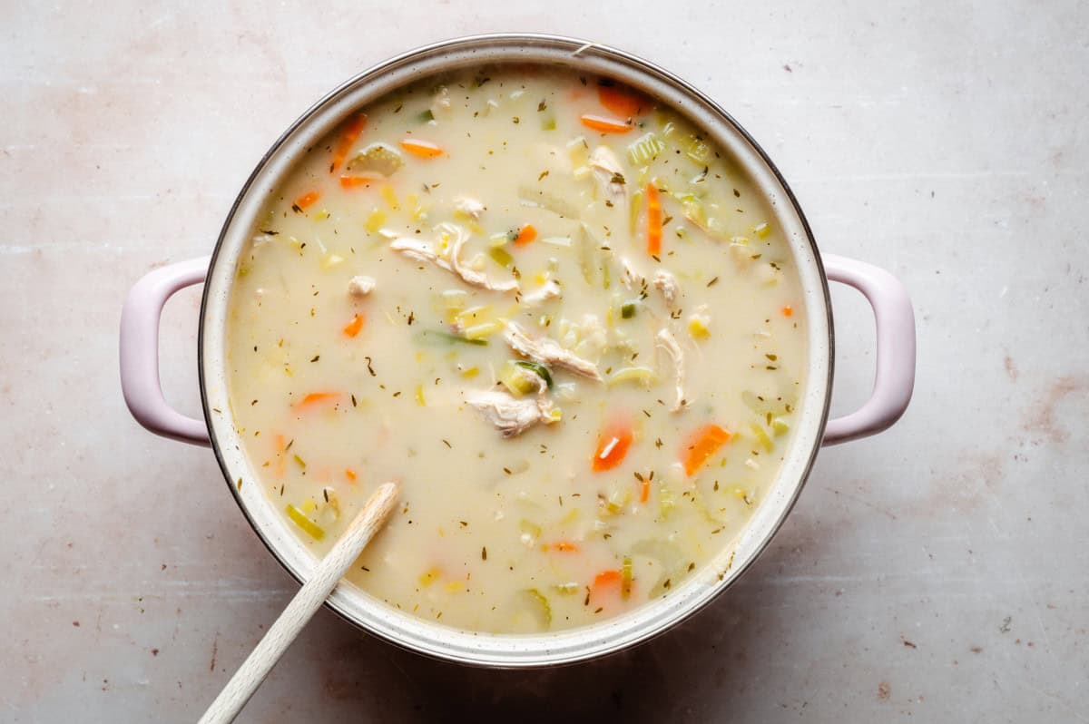 A top view of a pot filled with creamy chicken and vegetable soup, containing chunks of chicken, sliced carrots, celery, and herbs. A wooden spoon rests in the soup, and the pot has pale pink handles.