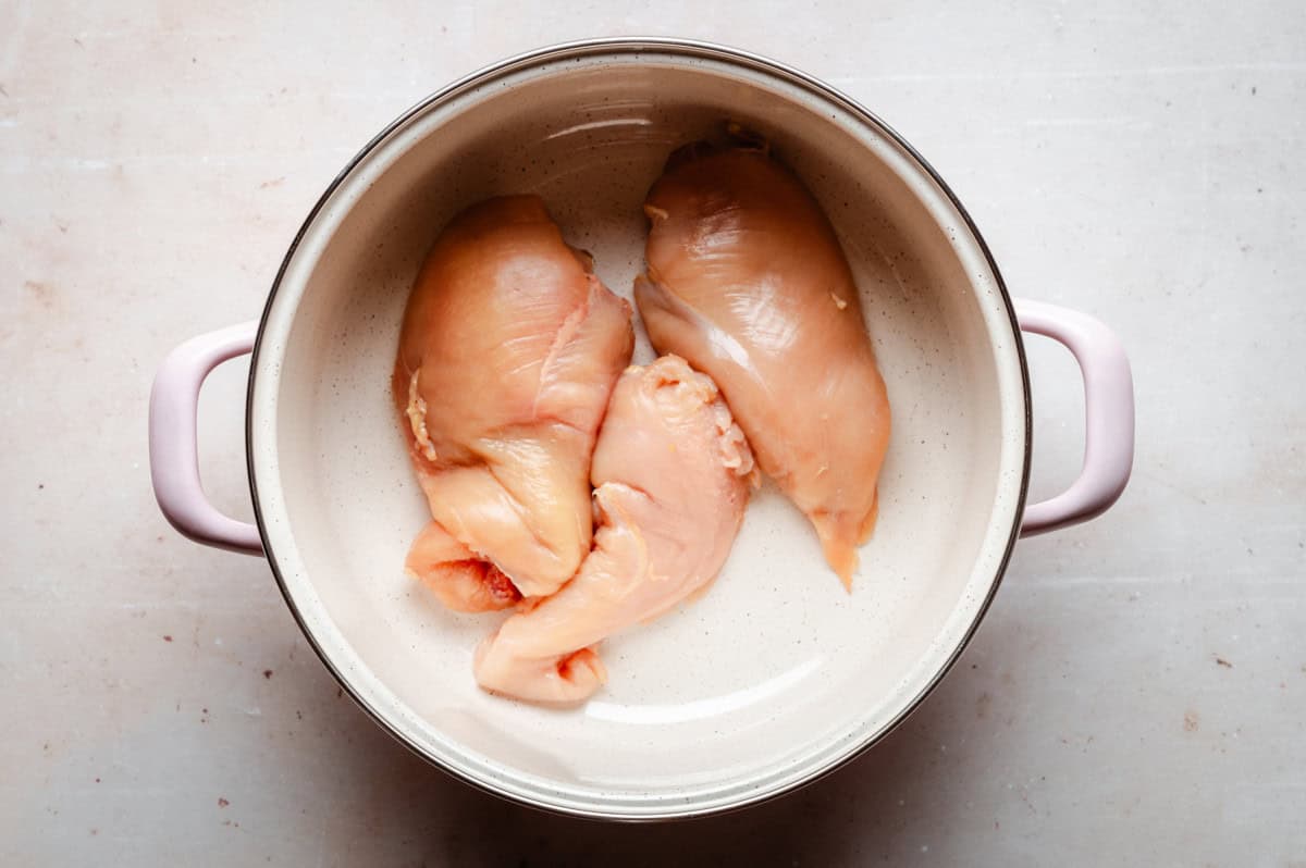 Three raw chicken breasts in a round, light-colored pot with pink handles, placed on a light-colored surface.