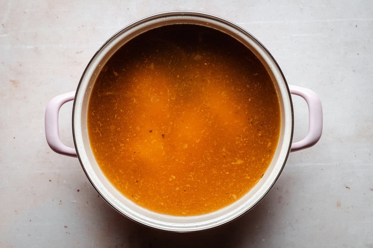 A top-down view of a pot filled with a golden-brown chicken broth. The pot has two handles on either side and rests on a pale countertop. The broth surface appears smooth with some visible particles.