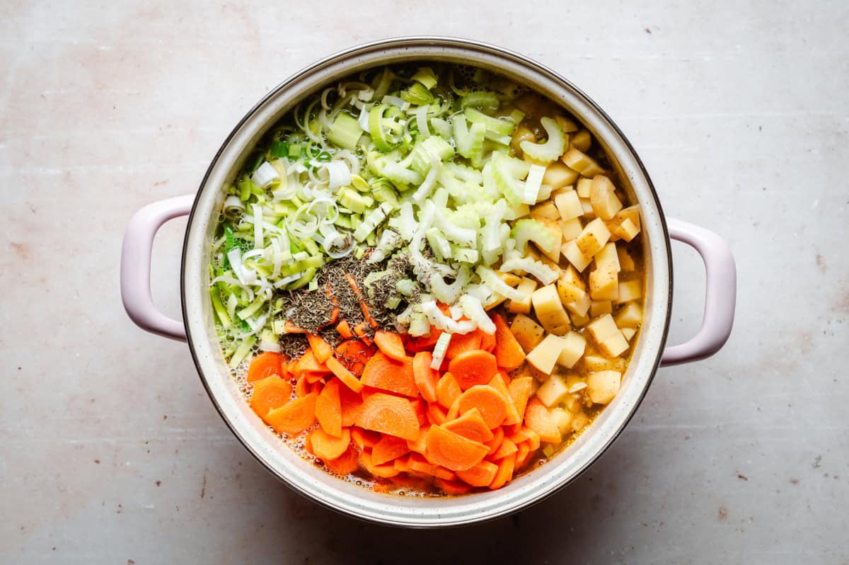 A pot filled with chopped vegetables, including carrots, celery, leeks, and potatoes, topped with herbs, ready for cooking. The pot sits on a light-colored surface.