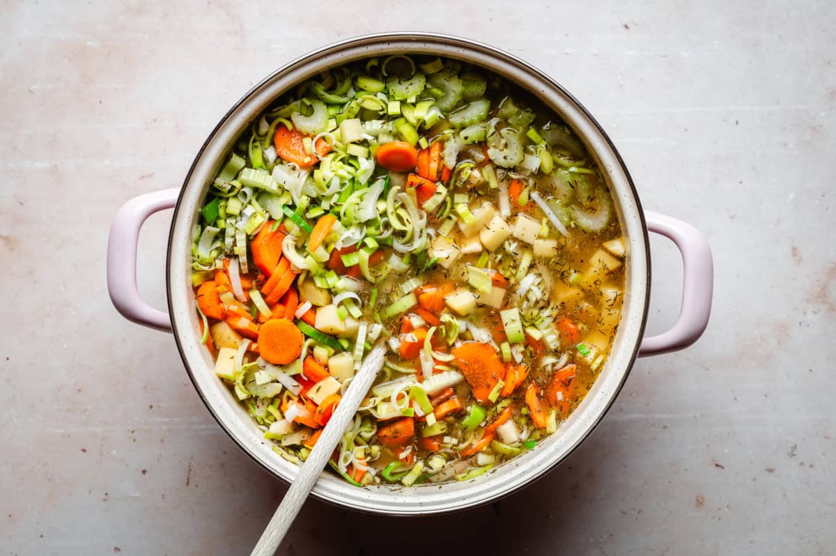 A pot of vegetable soup being cooked, containing sliced carrots, leeks, celery, and potatoes in a clear broth. A wooden spoon is placed in the pot. The pot has light pink handles and is resting on a light-colored surface.