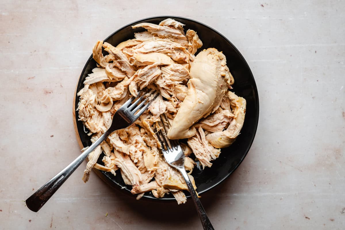 A black bowl filled with shredded chicken on a light surface. Two forks are positioned in the bowl, suggesting the chicken is being shredded or served.