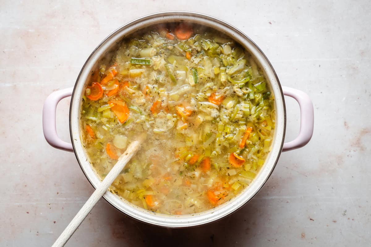 A large pot filled with a hearty vegetable soup, featuring visible pieces of carrots, celery, and herbs. A wooden spoon rests in the pot, and steam rises, indicating the soup is hot. The pot is set on a light-colored surface.