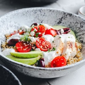 A ceramic bowl filled with a vibrant quinoa salad, topped with sliced baked chicken, avocado, cherry tomatoes, black olives, feta cheese crumbles, and a drizzle of creamy dressing. The dish is garnished with chopped herbs.