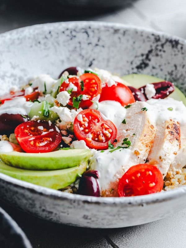 A ceramic bowl filled with a vibrant quinoa salad, topped with sliced baked chicken, avocado, cherry tomatoes, black olives, feta cheese crumbles, and a drizzle of creamy dressing. The dish is garnished with chopped herbs.