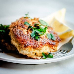 A close-up of a breaded and fried cod fish cake garnished with fresh parsley, served on a white plate. A lemon wedge can be seen in the background. A fork partially rests on the plate near the cod fish cake.