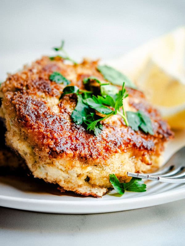 A close-up of a breaded and fried cod fish cake garnished with fresh parsley, served on a white plate. A lemon wedge can be seen in the background. A fork partially rests on the plate near the cod fish cake.