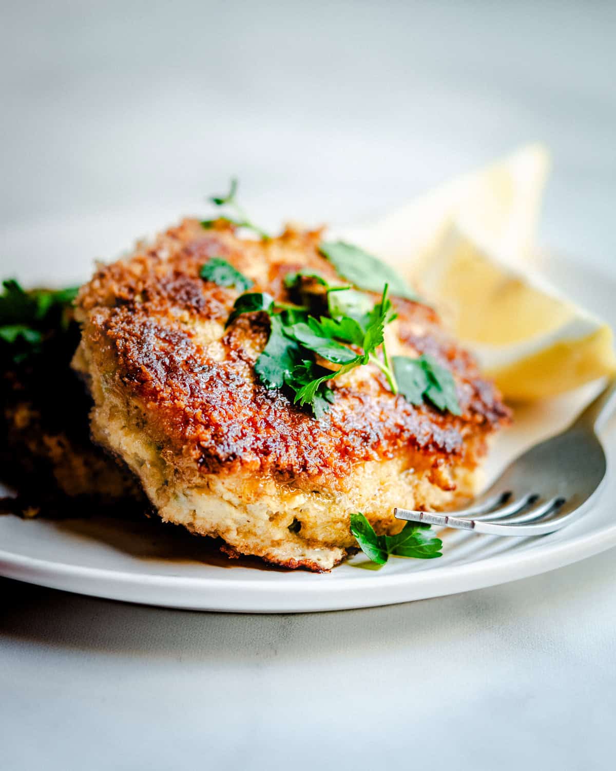 Close-up of a plate with a breaded and fried fish cake garnished with fresh parsley. A wedge of lemon is placed beside the fish cake, and a fork rests on the plate, ready to be used. The background is blurred, focusing attention on the dish.