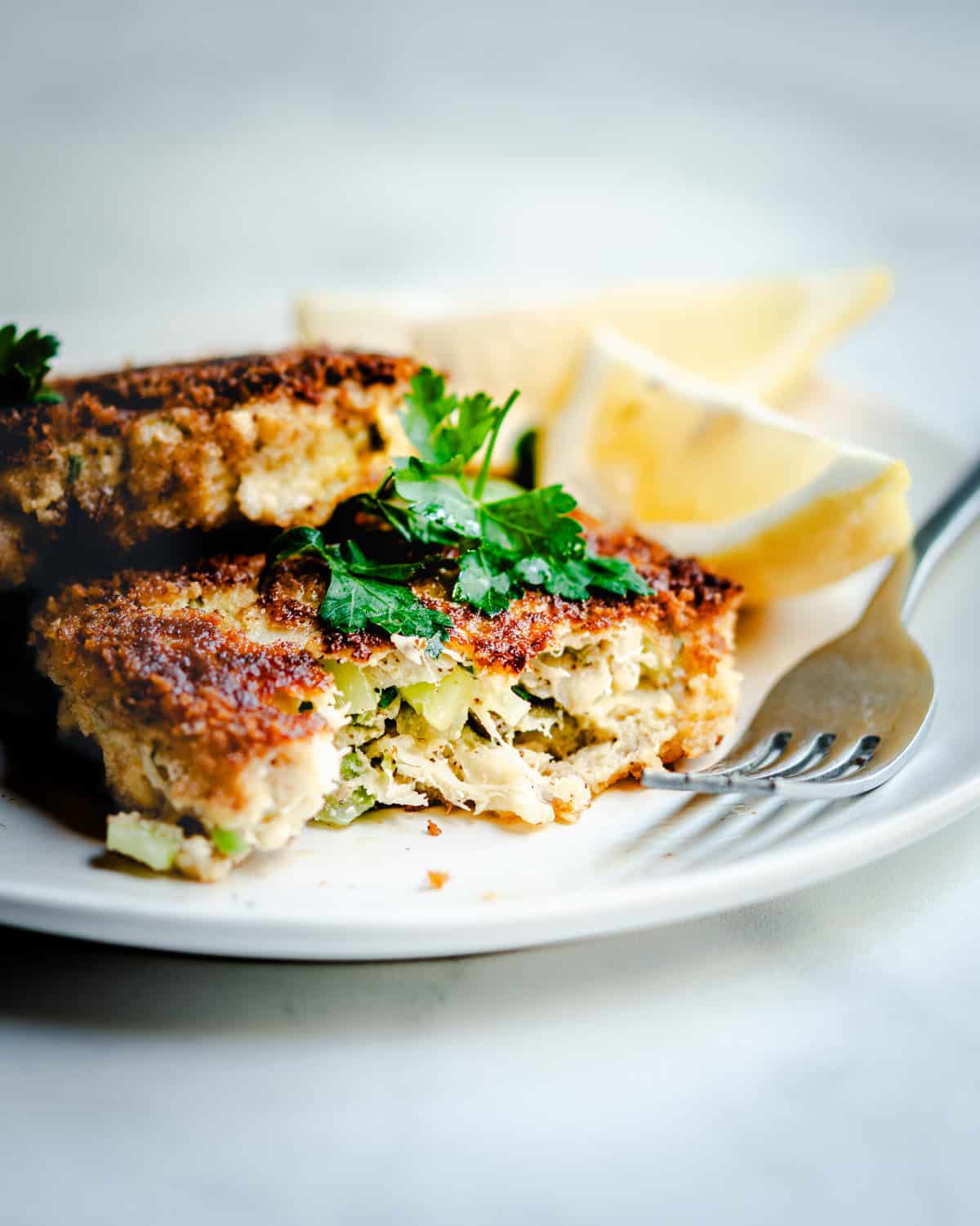 A close-up of a crispy, golden-brown cod fish cake garnished with fresh parsley and served with lemon wedges on a white plate. A fork is placed beside the fish cake, and the dish is set against a light background.