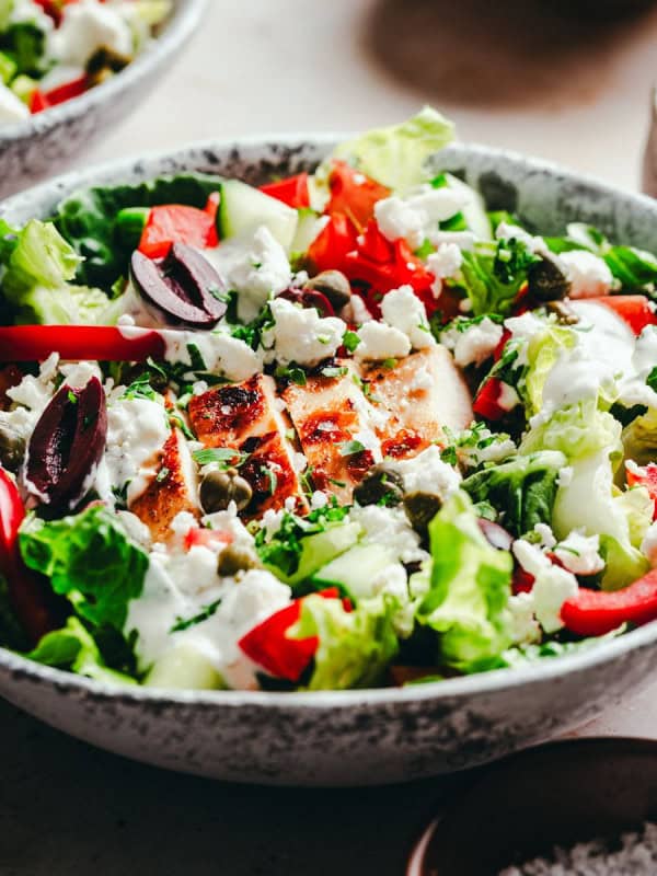 A bowl filled with a colorful Greek Chicken salad, including greens, sliced red bell peppers, cucumbers, olives, capers, and crumbled feta cheese. Tender chicken slices are mixed in, and a creamy dressing is drizzled on top. A fork rests on the side of the bowl.