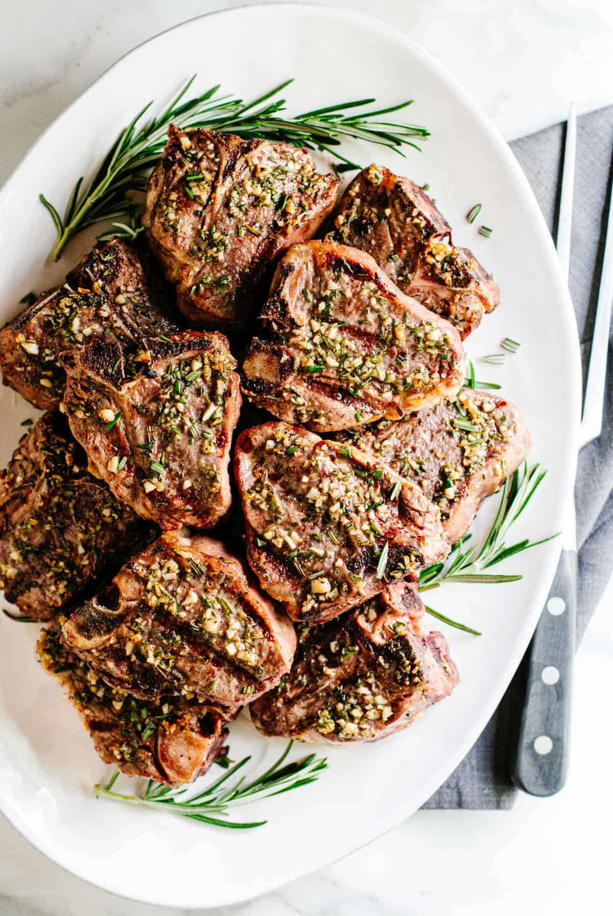 Grilled Lamb Loin Chops stacked on a white platter with rosemary sprigs photographed from above.