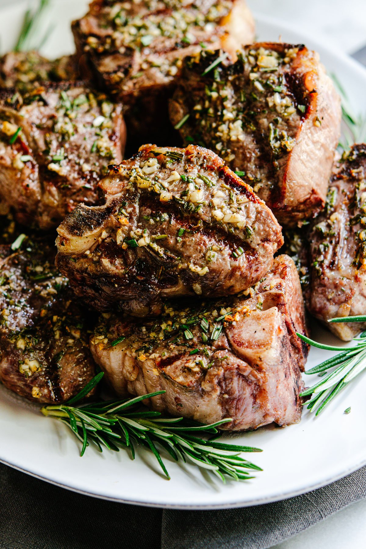 Grilled Lamb Loin Chops stacked on a white platter with rosemary sprigs.