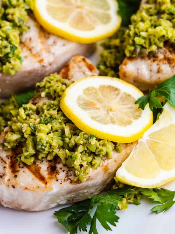 Grilled Swordfish served on a platter and topped with green olive tapenade, lemon slices and parsley.