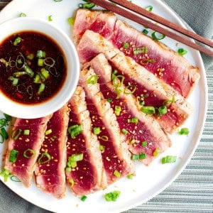 A plate of grilled tuna slices garnished with chopped green onions and a sprinkle of seasoning. There is a small bowl of dipping sauce with sliced green onions on the side. Chopsticks rest on the edge of the plate. The background includes a textured cloth and mat.