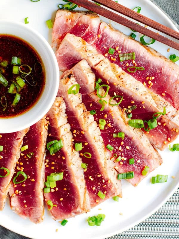 A plate of grilled tuna slices garnished with chopped green onions and a sprinkle of seasoning. There is a small bowl of dipping sauce with sliced green onions on the side. Chopsticks rest on the edge of the plate. The background includes a textured cloth and mat.