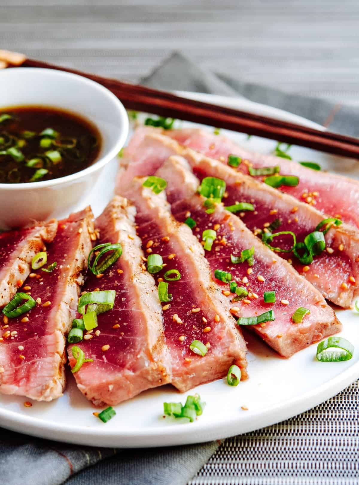 A white plate features neatly sliced pieces of seared tuna, garnished with chopped scallions. A small bowl of soy-based dipping sauce sits beside the tuna, accompanied by a pair of wooden chopsticks resting on the edge of the plate.