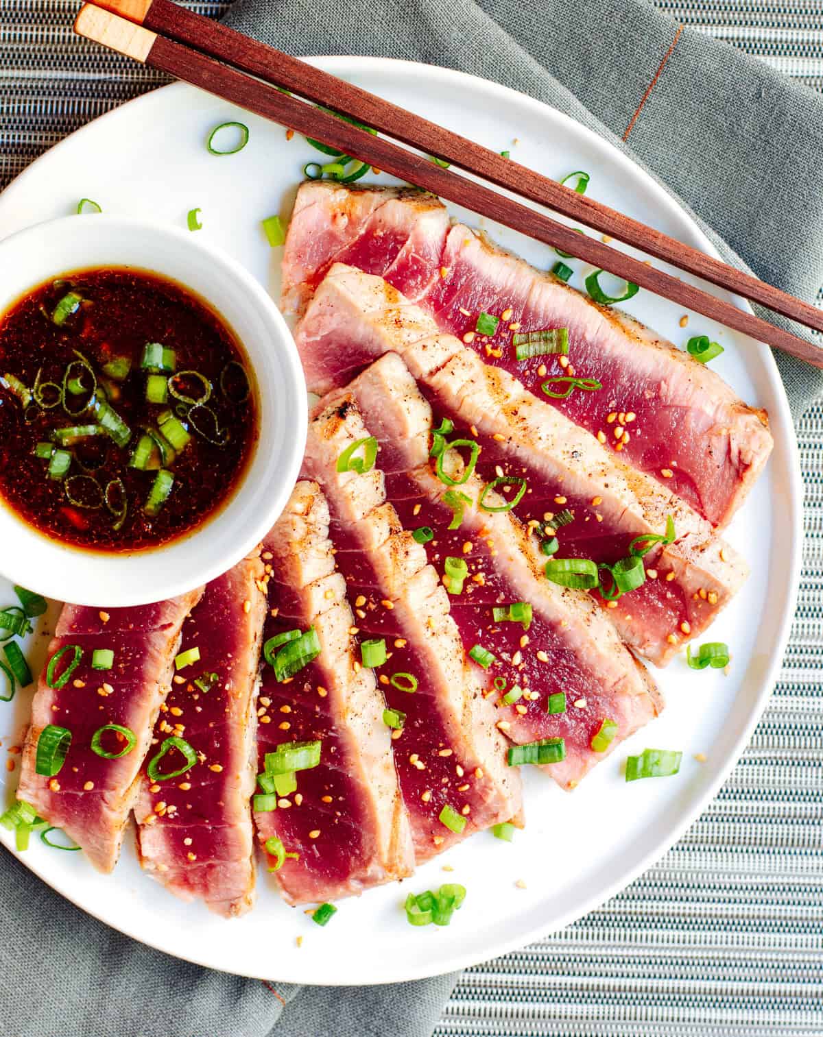 A plate of seared tuna slices garnished with chopped green onions and sesame seeds, accompanied by a small bowl of soy-based dipping sauce. Chopsticks rest on the edge of the plate. The plate is set on a blue-gray placemat.