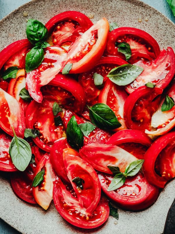 A plate of fresh tomato salad topped with basil leaves and sprinkled with salt. The salad is arranged on a rustic stone plate with a fork placed on the side. The vibrant red of the tomatoes contrasts with the green of the basil, creating a visually appealing dish.