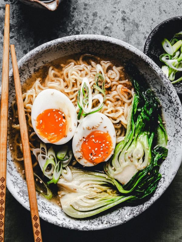 A bowl of ramen with sliced boiled eggs, bok choy, and green onions. Chopsticks rest on the bowl's edge. A small dish with more green onions is beside the bowl on a textured gray surface.