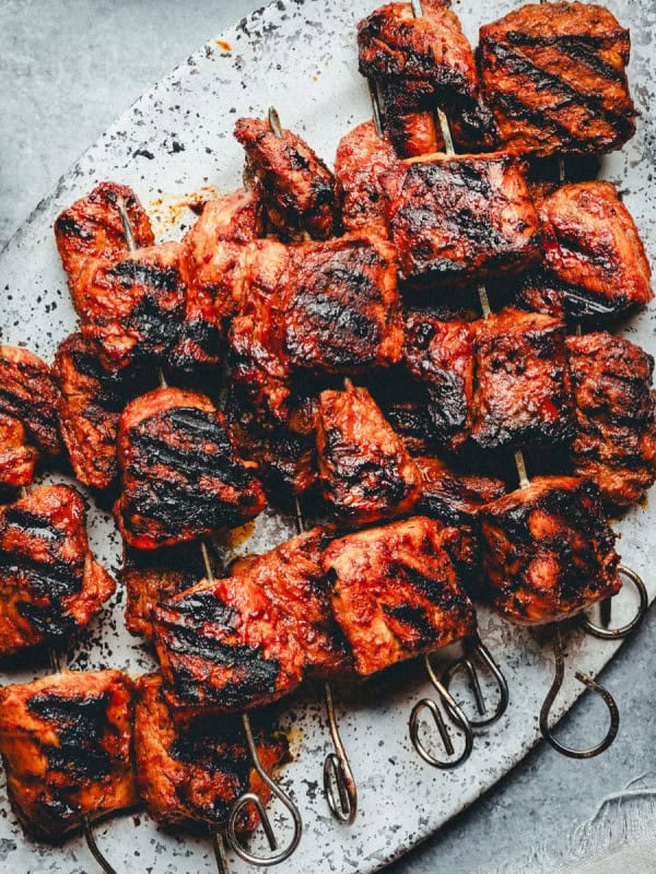 A rustic metal tray holds several skewers of grilled pork, charred and juicy. Visible grill marks enhance the rich, reddish-brown color of the cooked meat. To the side is a spoonful of bright red sauce, and a dark beverage in a glass. The background is a textured grey.