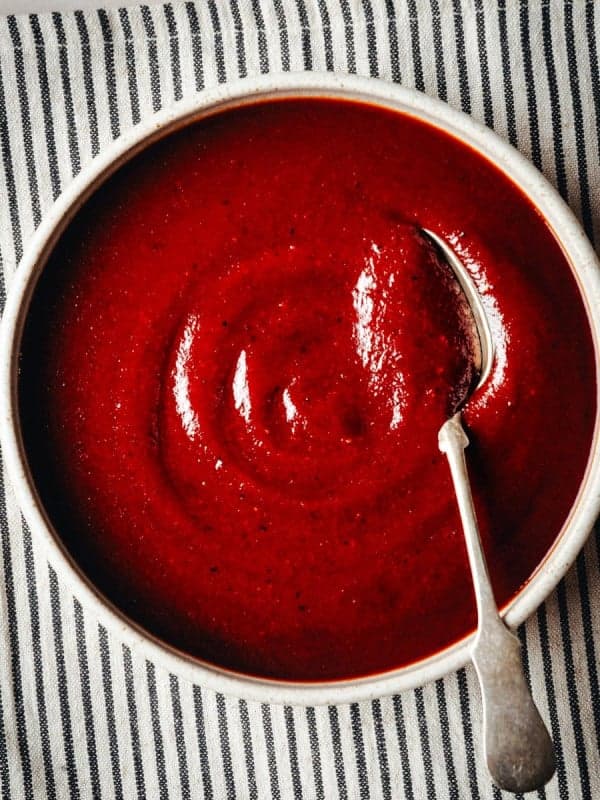 A bowl of smooth, red BBQ Sauce sits on a striped black-and-white cloth. A silver spoon rests on the edge of the bowl, its handle pointing outward. The vibrant color of the soup contrasts with the neutral tones of the cloth and table.